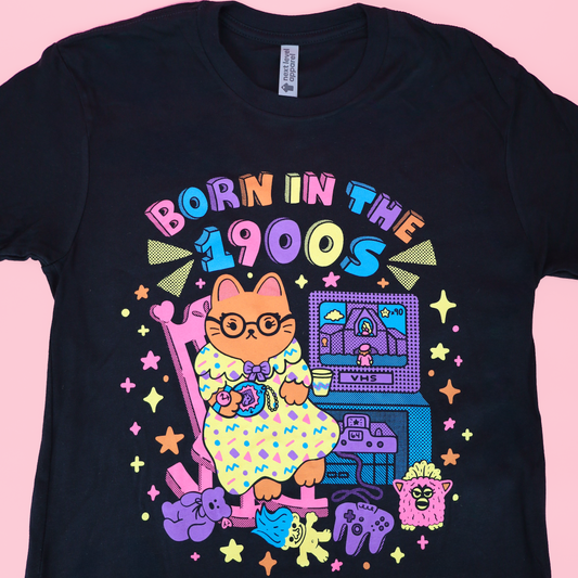 A black short-sleeve graphic t-shirt featuring a colorful retro-style illustration of a cat in a polka-dot dress playing a keyboard, with the text 'BORN IN THE 1900S' above it, shown against a solid pink background.