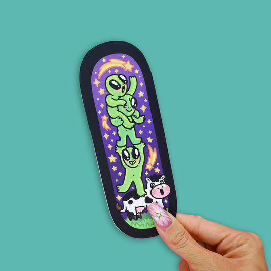 A black-bordered bookmark with a colorful cartoon illustration of green aliens stacking on a cow, placed on an open book against a teal background.