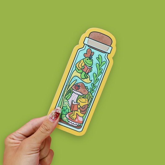 A die-cut sticker shaped like a glass bottle with cartoon frogs reading under a mushroom, placed on an open book against a bright green background.