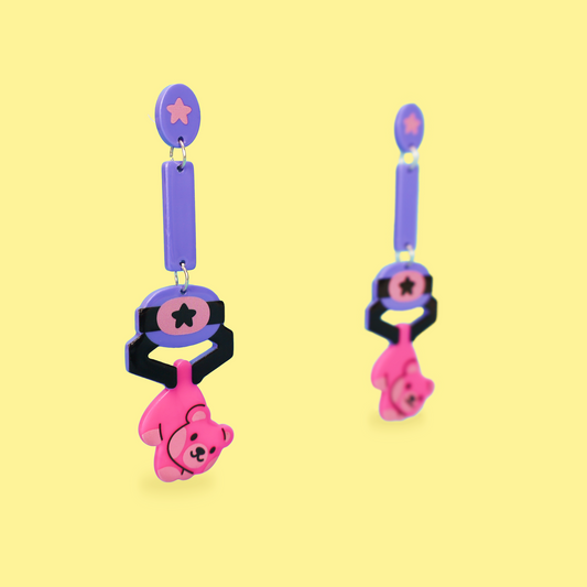 A pair of dangling earrings on a pale yellow background. Each earring has a lavender oval stud with a pink star, a lavender rectangular drop, a black and pink circular section with a star, and a dangling pink bear charm with black facial features.