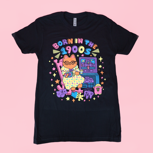 A black short-sleeve graphic t-shirt featuring a colorful retro-style illustration of a cat in a polka-dot dress playing a keyboard, with the text 'BORN IN THE 1900S' above it, shown against a solid pink background.