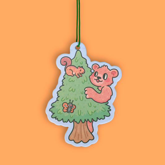 A die-cut hanging ornament featuring a cartoon pine tree with a pink bear climbing it, a squirrel on the tree, and a butterfly, suspended by a thin green string against a solid orange background.