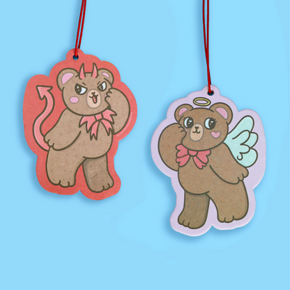 Two cartoon bear-shaped hanging tags or ornaments on a light blue background. One bear has devil horns, a pink bow, and a red outline with a red hanging string.