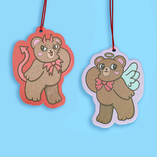 Two cartoon bear-shaped hanging tags or ornaments on a light blue background. One bear has devil horns, a pink bow, and a red outline with a red hanging string.