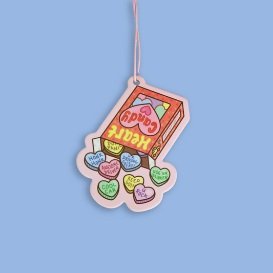 A novelty car air freshener or ornament shaped like a box of Heart Candy, featuring colorful heart-shaped candies spilling out, suspended by a thin pink string against a solid light blue background.
