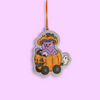 A die-cut Halloween ornament featuring a purple cat in an orange pumpkin car with a witch hat, hanging by an orange string against a solid light purple background.