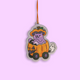 A die-cut Halloween ornament featuring a purple cat in an orange pumpkin car with a witch hat, hanging by an orange string against a solid light purple background.