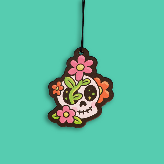 A colorful sugar skull hanging ornament with pink and orange flowers, green leaves, and black details, suspended by a black string against a solid teal background.