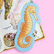 A die-cut paper bookmark shaped like a seahorse, featuring a light blue background with an illustrated orange and yellow seahorse. The bookmark is layered over an open book with seashell illustrations and is photographed against a solid pink background.