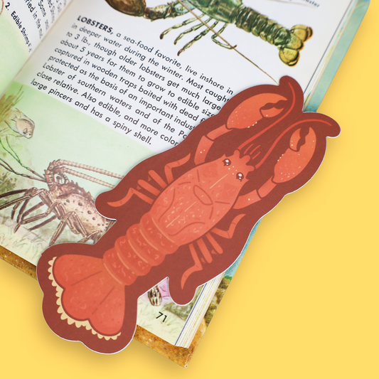 A lobster-shaped bookmark made of layered paper in reddish-brown and orange tones, placed on an open book with sea life illustrations, against a solid yellow background.