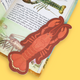 A lobster-shaped bookmark made of layered paper in reddish-brown and orange tones, placed on an open book with sea life illustrations, against a solid yellow background.
