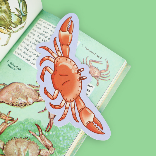 A die-cut sticker shaped like a cartoon orange crab bookmarking an open children's book with illustrations, set against a solid light green background.