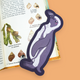 A die-cut sticker shaped like a cartoon penguin in shades of purple and gray, placed on an open book with shell illustrations against a solid peach background.