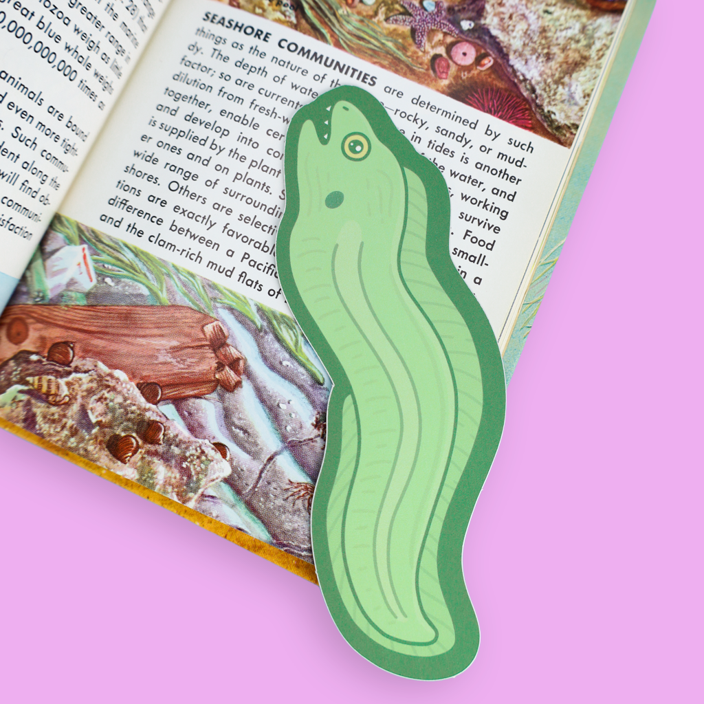 A green cucumber-shaped paper bookmark with a die-cut outline, placed on an open book with a light pink background.
