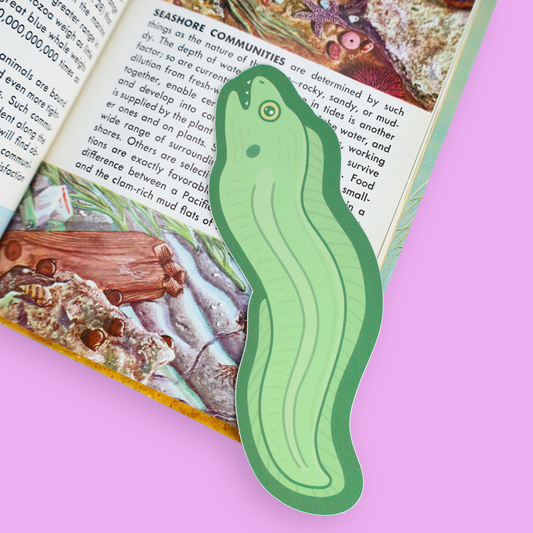 A green cucumber-shaped paper bookmark with a die-cut outline, placed on an open book with a light pink background.