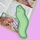 A green cucumber-shaped paper bookmark with a die-cut outline, placed on an open book with a light pink background.