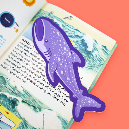 A purple shark-shaped bookmark with white speckles placed on an open children's science book about waves, set against a solid coral background.