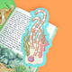 A die-cut educational book plate featuring an underwater scene with striped coral, green seaweed, a purple clam shell, and a blue border, shown resting on an open book against an orange background.