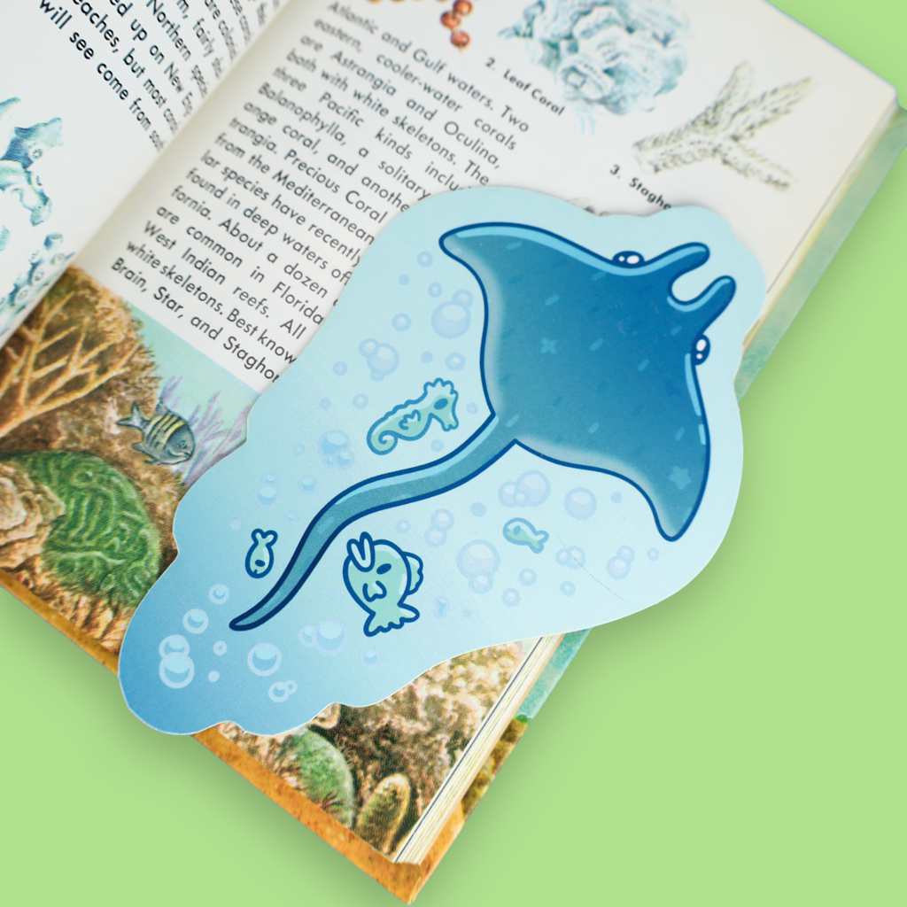 A light blue and blue-gray magnetic bookmark shaped like a manta ray with sea creatures, shown resting on an open illustrated book against a light green background.