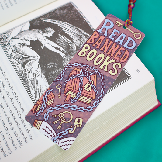 A decorative bookmark shaped like a book cover with the phrase “READ BANNED BOOKS” in large pastel letters, featuring illustrations of stacked books wrapped in chains and keys. The bookmark is placed over an open book on a bright teal background.
