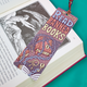 A decorative bookmark shaped like a book cover with the phrase “READ BANNED BOOKS” in large pastel letters, featuring illustrations of stacked books wrapped in chains and keys. The bookmark is placed over an open book on a bright teal background.