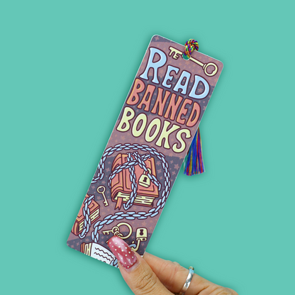 A hand with pink glitter nail polish holds a rectangular bookmark featuring the text 'READ BANNED BOOKS' in stylized lettering, with illustrations of books, keys, and rope on a purple background. The bookmark has a small tassel at the top and is photographed against a solid teal background.