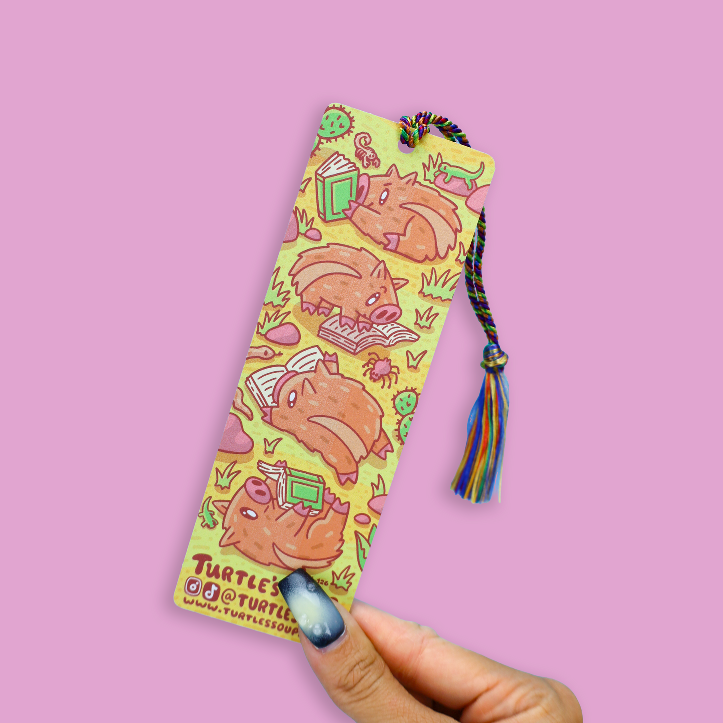 A hand with blue nail polish holds a long, narrow yellow bookmark featuring cartoon turtle illustrations and a multicolored tassel, against a solid pink background.