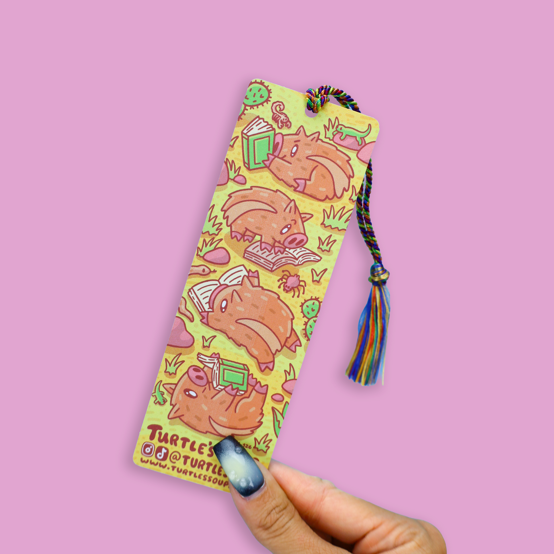 A hand with blue nail polish holds a long, narrow yellow bookmark featuring cartoon turtle illustrations and a multicolored tassel, against a solid pink background.