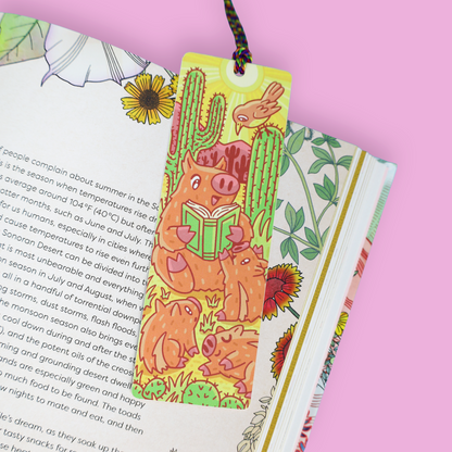 A colorful illustrated bookmark with a pig reading a book and a cactus design, hanging from a string between two open books on a pink background.