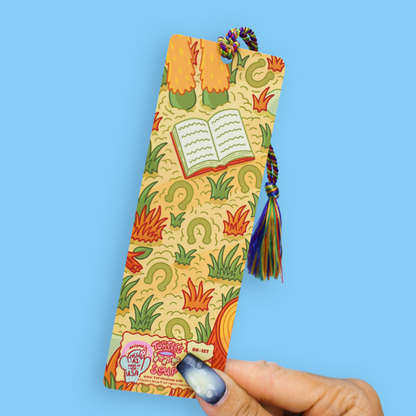 A hand holds a decorative paper bookmark featuring a colorful illustration of legs, an open book, and flames on a yellow background, with a braided tassel. The hand has blue and white galaxy nail art against a solid light blue background.
