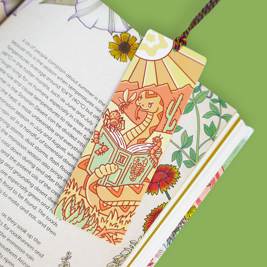 A decorative illustrated bookmark with a desert dragon reading in a library, attached to a book with patterned pages on a solid green background.