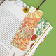A decorative illustrated bookmark with a desert dragon reading in a library, attached to a book with patterned pages on a solid green background.