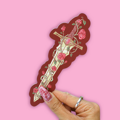 A hand with pink glitter nail polish holds a die-cut sticker of a decorative sword with pink roses and green leaves, set against a solid pink background.
