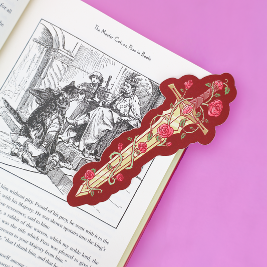 A decorative red and gold bookmark shaped like a sword with pink roses, placed on an open book page against a bright pink background.
