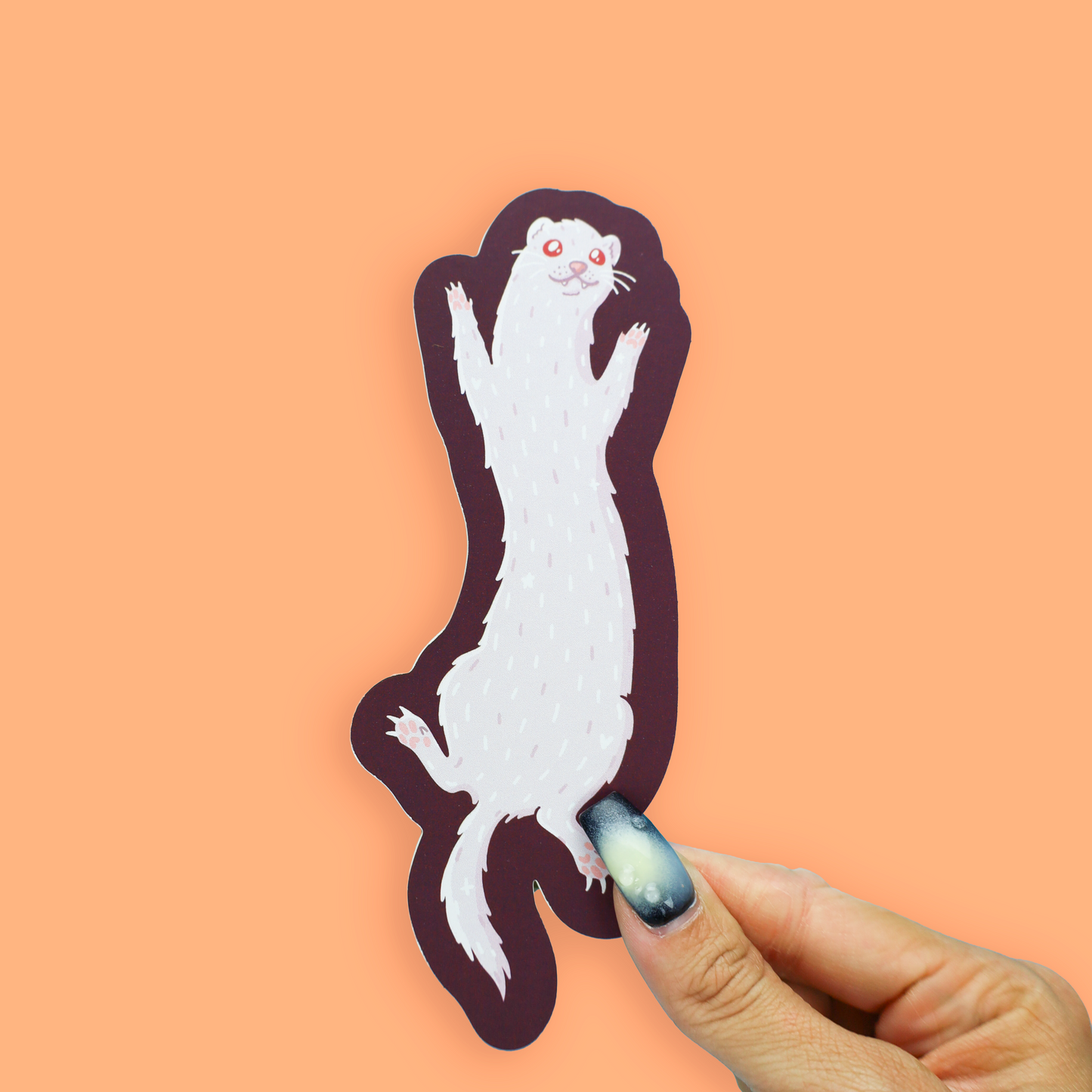 A die-cut sticker of a white ferret with red eyes and a dark plum outline, held by a hand with a galaxy-themed nail polish against a solid peach background.
