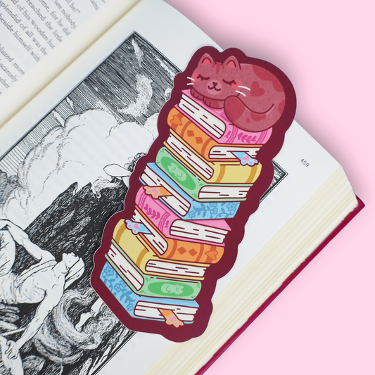 A die-cut sticker featuring a sleeping pink cat curled up on a stack of colorful illustrated books, placed on an open book against a light pink background.