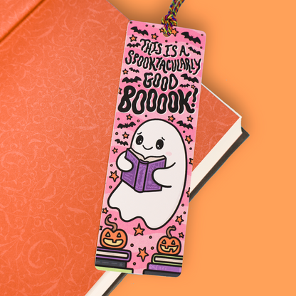 A pink Halloween-themed bookmark with a cute cartoon ghost reading a purple book, surrounded by stars, bats, pumpkins, and the text 'THIS IS A SPOOKTACULARLY GOOD BOOOOOOK!', shown resting on an orange notebook against a solid orange background.