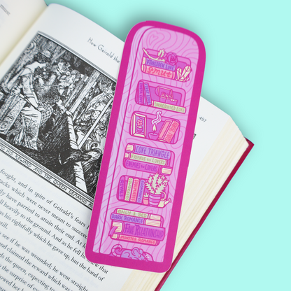 A pink rectangular book bookmark with illustrated book spines and book-themed text, placed on an open book against a mint blue background.