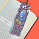 A colorful illustrated bookmark shaped like a tree trunk and birdhouse, placed across an open book on an orange background. The bookmark features a blue bird holding a green bowl, with shelves, books, and small whimsical details.