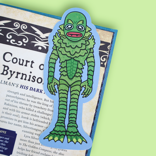 A die-cut sticker of a green scaly creature resembling the Lizard from The Lord of the Rings, placed on top of an open book page against a light green background.