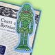 A die-cut sticker of a green scaly creature resembling the Lizard from The Lord of the Rings, placed on top of an open book page against a light green background.