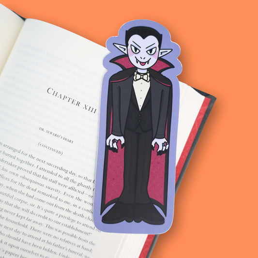 A vampire-shaped book bookmark with a blue border, featuring a cartoon vampire in a black and red cape, resting on an open book against an orange background.