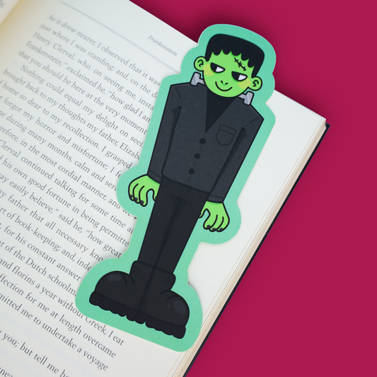 A die-cut paper bookmark featuring a cartoon stitched monster character in a black suit with green skin, placed on an open book with a solid pink background visible around it.