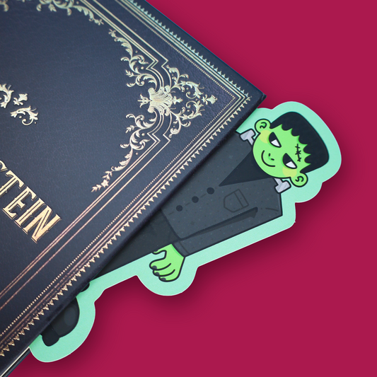 A die-cut paper bookmark featuring a cartoon stitched monster character in a black suit with green skin, placed on an open book with a solid pink background visible around it.