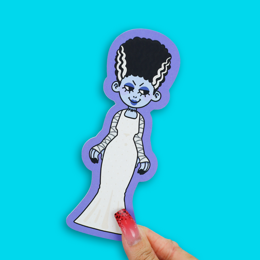 A die-cut sticker of a cartoon mummy character with black hair, a white dress, and a purple border, held by a hand with red glitter nail polish against a bright cyan background.