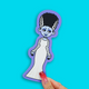 A die-cut sticker of a cartoon mummy character with black hair, a white dress, and a purple border, held by a hand with red glitter nail polish against a bright cyan background.