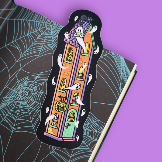 A Halloween-themed bookmark shaped like a haunted house, featuring cartoon ghosts and a purple cat, placed on an open book with a spiderweb pattern against a solid purple background.