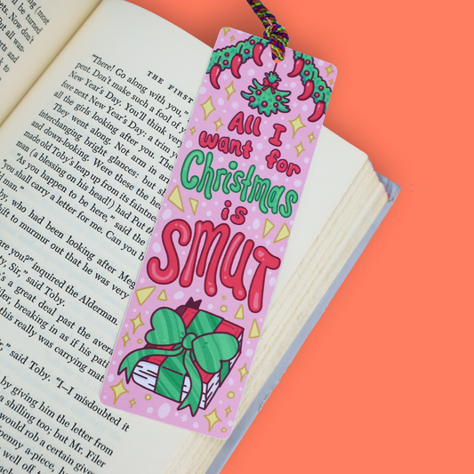 A pink Christmas-themed bookmark with the text 'All I want for Christmas is SMUT' in green and red lettering, decorated with illustrations of a Santa hat, a wrapped gift, and gold stars, resting on an open book against a solid orange background.