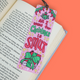 A pink Christmas-themed bookmark with the text 'All I want for Christmas is SMUT' in green and red lettering, decorated with illustrations of a Santa hat, a wrapped gift, and gold stars, resting on an open book against a solid orange background.