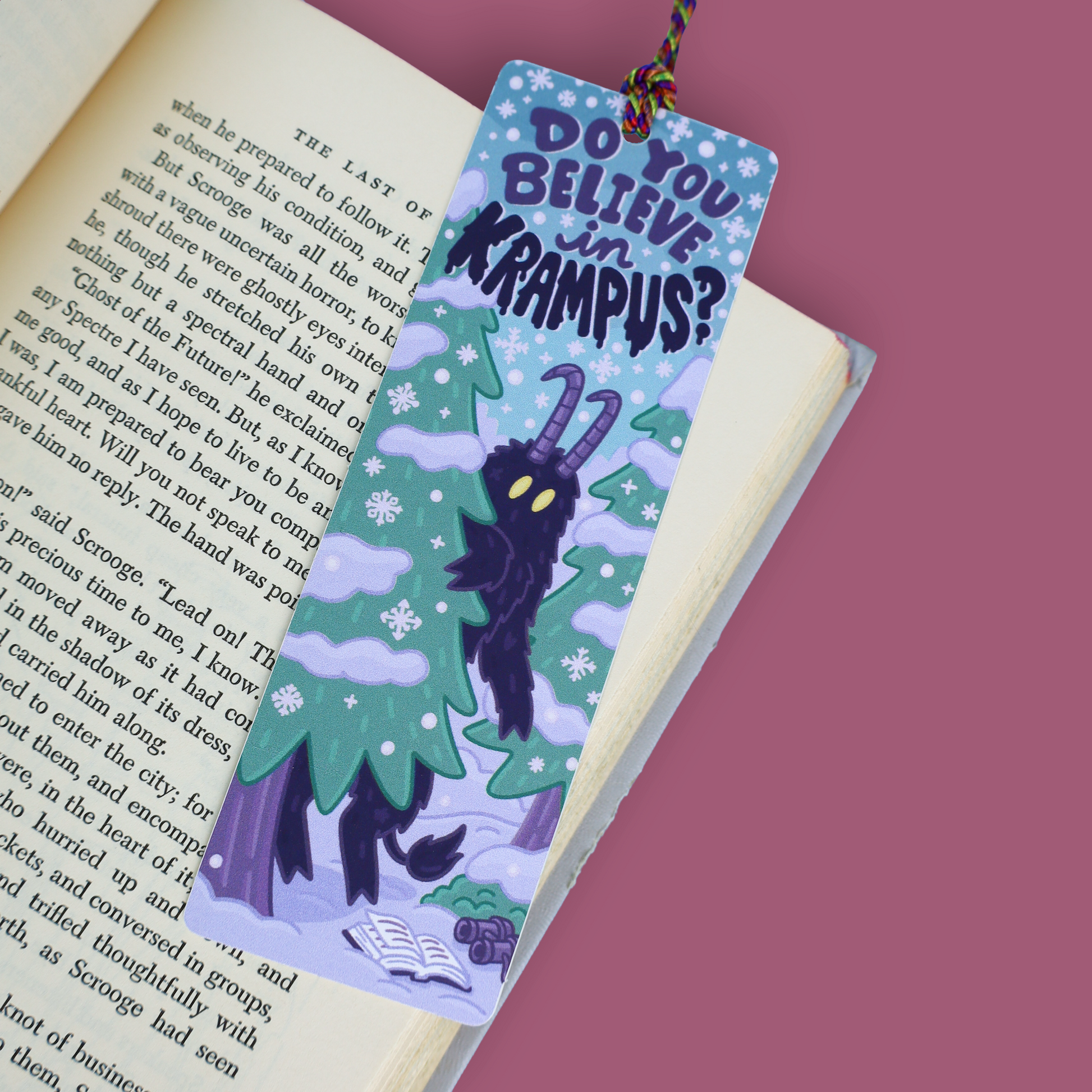 A decorative paper bookmark with a Krampus illustration and the text 'DO YOU BELIEVE IN KRAMPUS?' placed on an open book against a solid pink background.